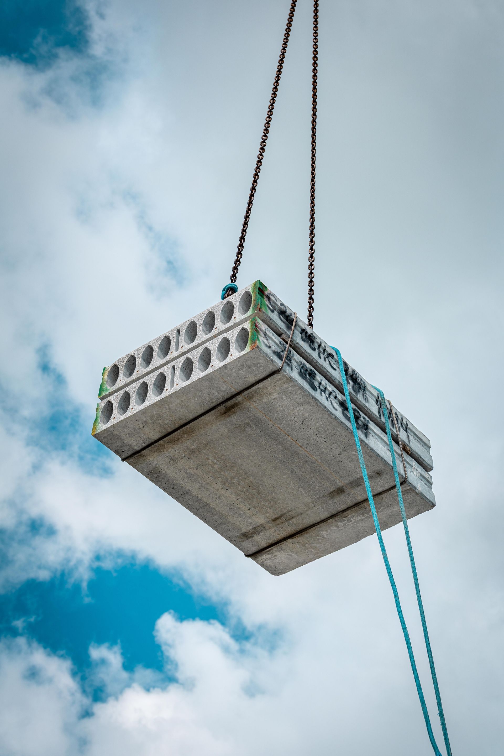 Concrete slab being lifted by crane cables against a cloudy blue sky