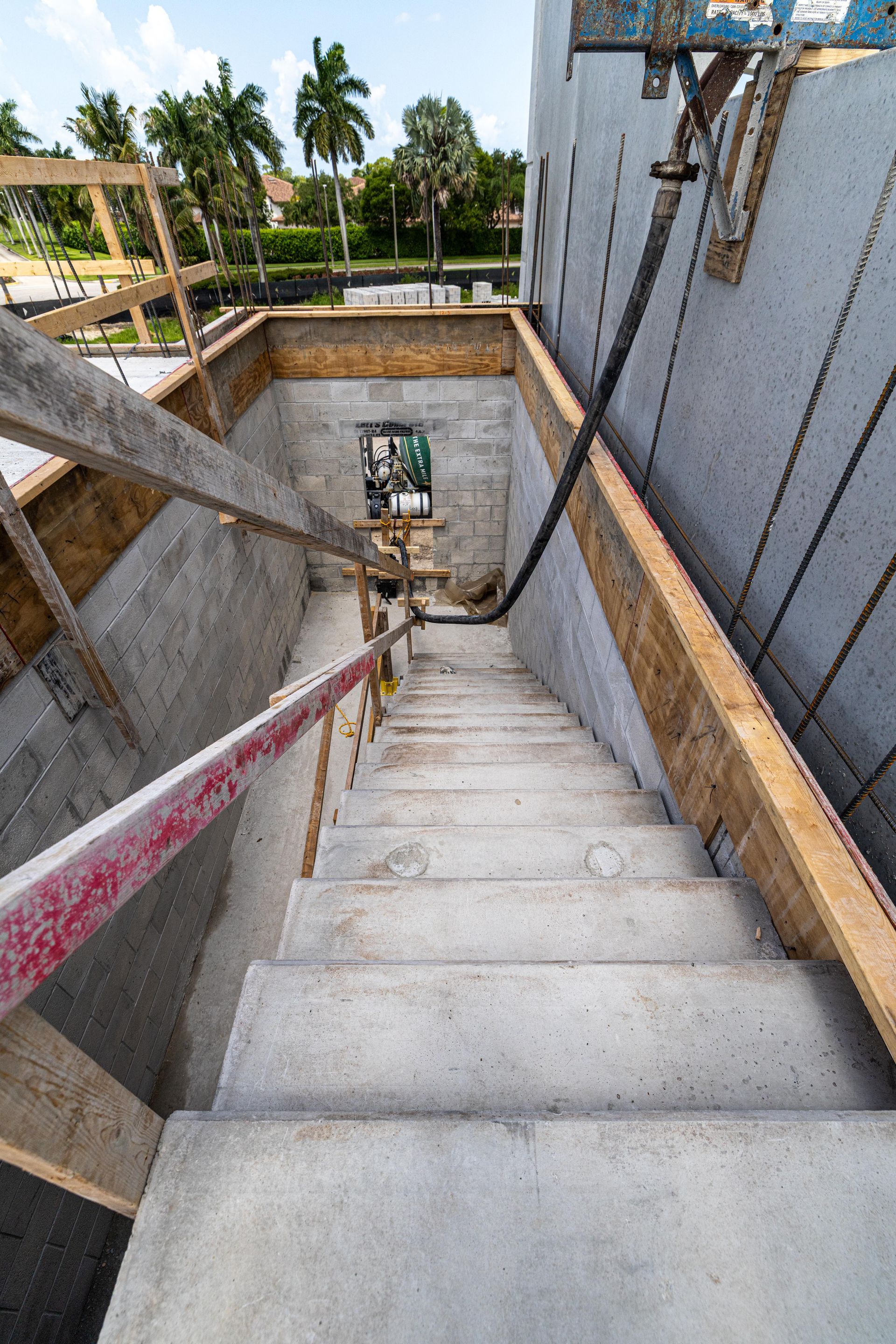 Steep concrete stairway in an unfinished building, with handrails and palm trees visible outside.
