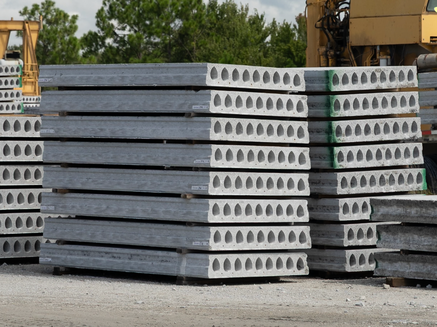 Stacks of white concrete slabs with oval holes in a gravel yard under a blue sky.