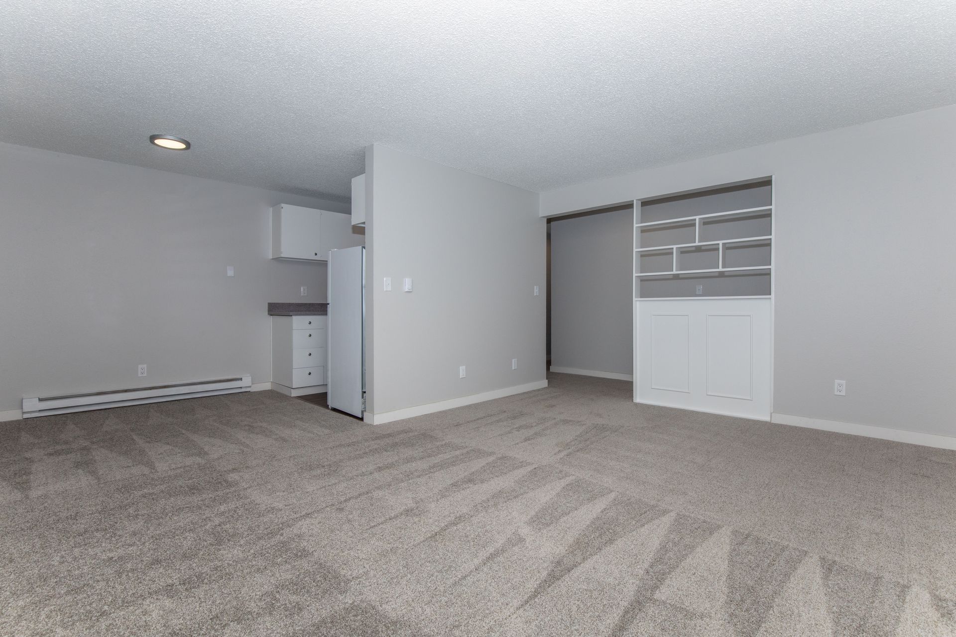 An empty living room with a carpeted floor and a refrigerator.