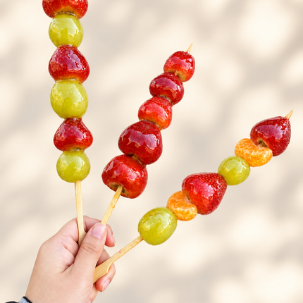 Hand holding three skewers of glossy red and yellow candied fruit against a soft background