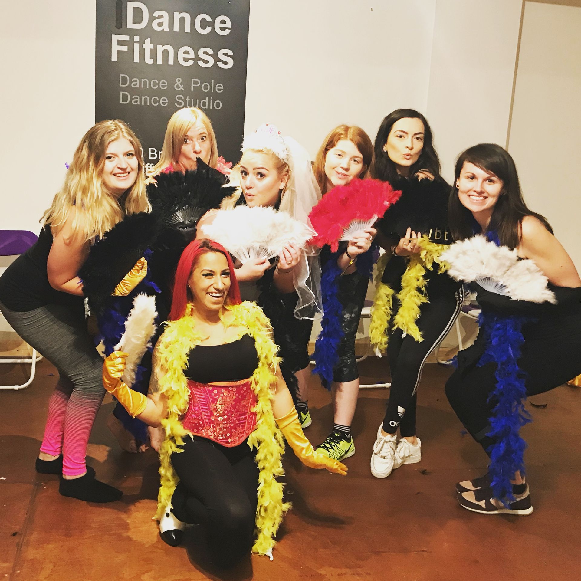 Group of women posing with feather boas and fans at a burlesque themed hen party dance class, dressed in glamorous outfits in a studio