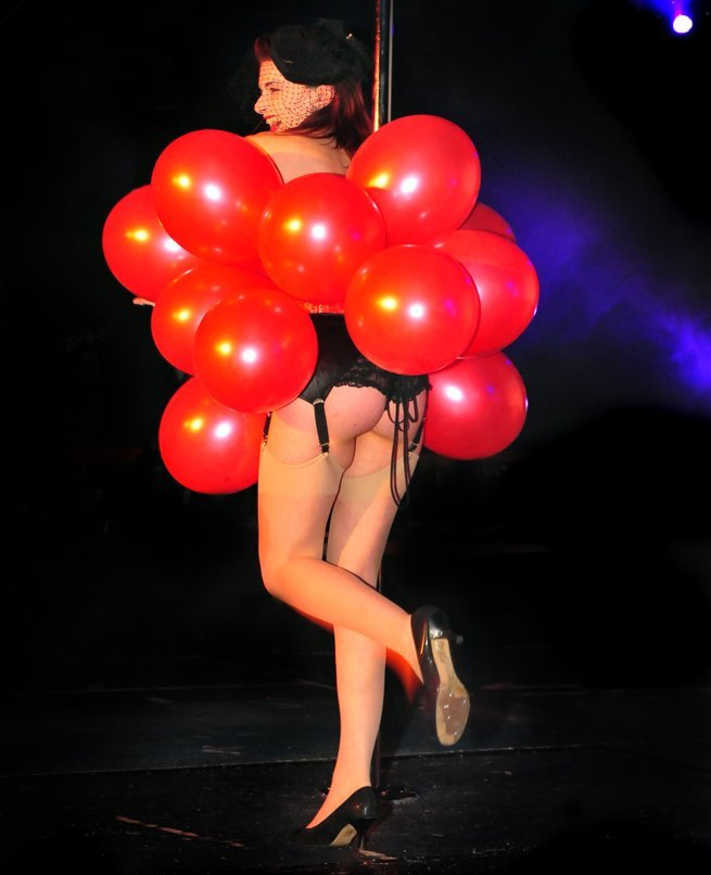 Burlesque dancer dressed in a red balloon-themed costume performing on stage with dramatic spotlight