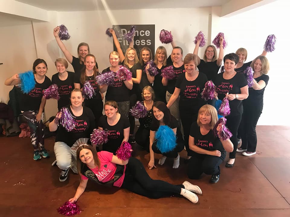 Group of women holding pom poms at a cheerleading themed hen party dance class, posing together in a studio wearing matching outfits