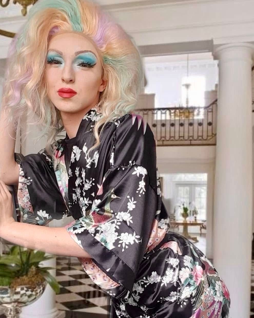 Elegant portrait of drag queen DB in Poole wearing a pastel rainbow wig, glamorous makeup and a black floral kimono robe while posing inside a luxurious grand interior setting.