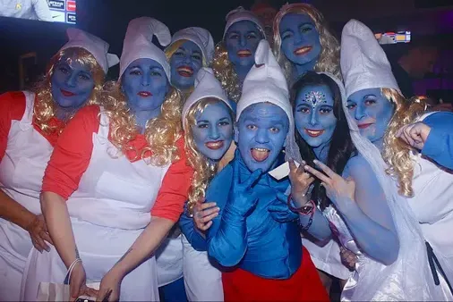 Group of hens and male dwarf all dressed as Smurfs in blue face paint and themed costumes, posing for photos at a fancy dress party and themed event.