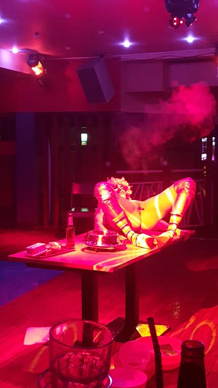 London fetish performer reclining on a table under vivid pink stage lighting during an immersive fetish cabaret show. The performer wears themed costume accessories and knee-high footwear, creating a dramatic alternative entertainment experience.