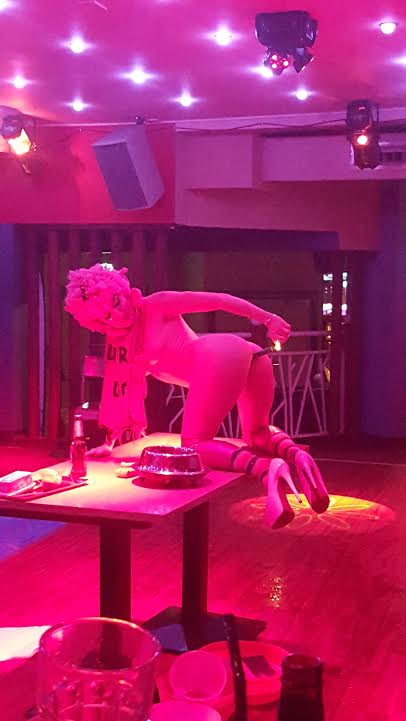 London fetish performer on stage under pink lighting, kneeling on a table during an immersive fetish cabaret performance. The performer wears high heels, themed accessories and theatrical makeup as part of an alternative entertainment show.