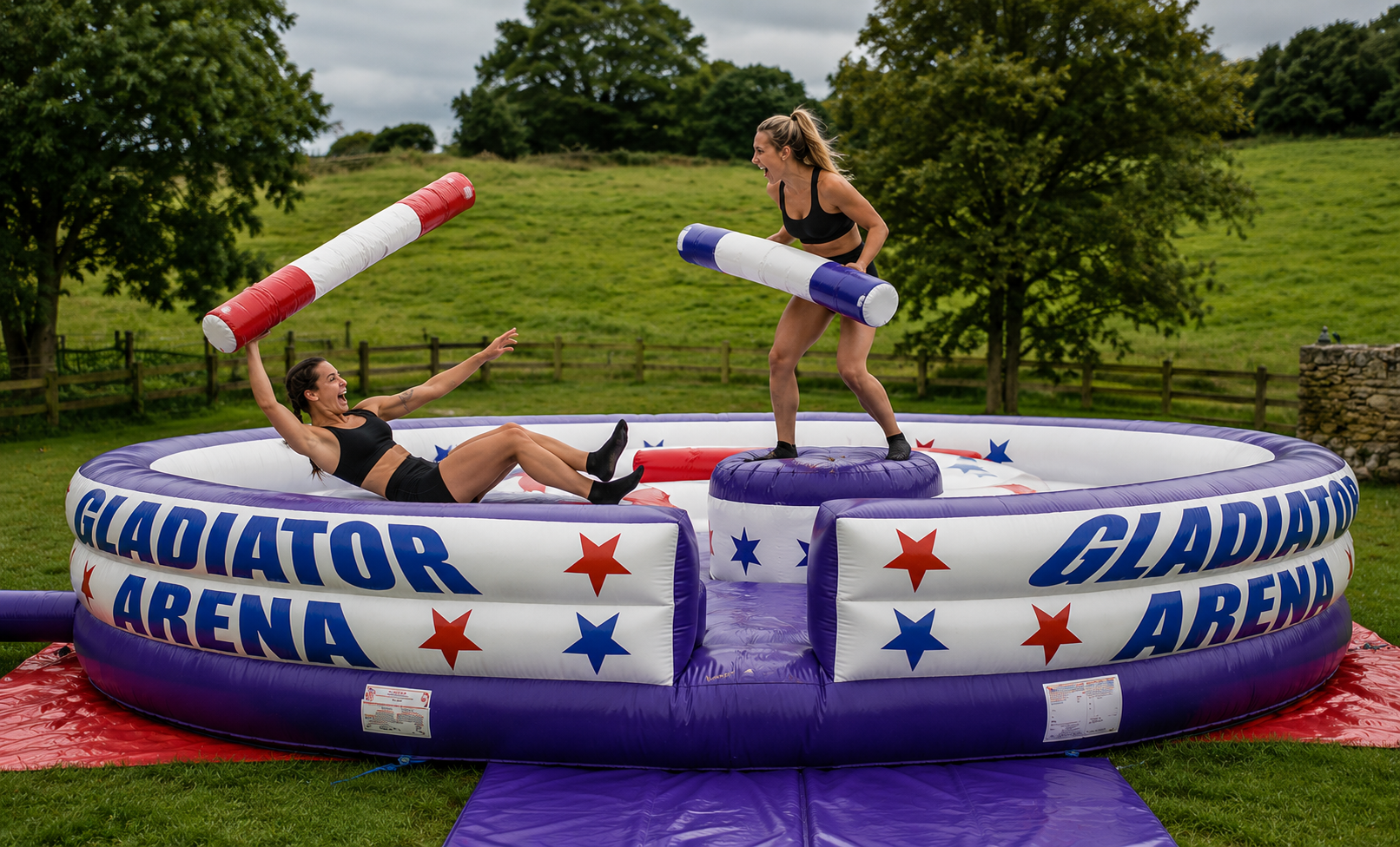 Two female gladiators battling with padded pugil sticks in a large inflatable gladiator arena at an outdoor party.