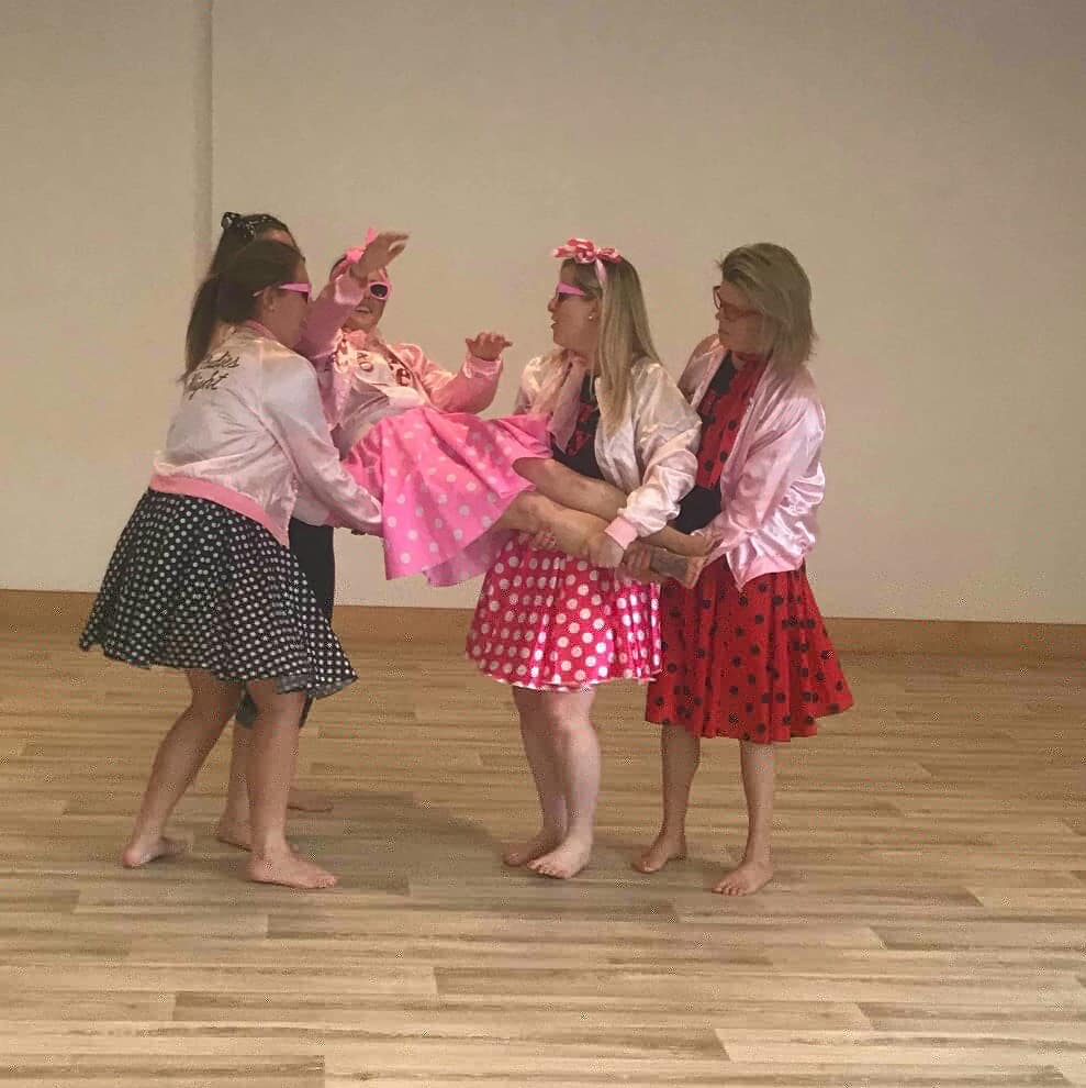 Group of women dressed as Grease Pink Ladies lifting a friend at a hen party dance class, wearing retro 50s outfits in a studio