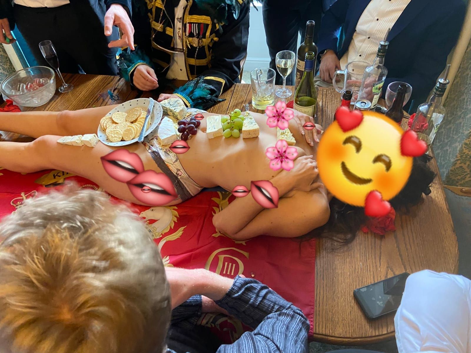 A glamorous human cheeseboard experience at a stag party, with a female food platter model lying across a wooden table while an elegant selection of cheeses, grapes, crackers and charcuterie accompaniments are beautifully arranged on her body. Smartly dressed stag party guests in suits and themed costumes gather around enjoying the interactive dining experience, creating a fun, sophisticated and memorable party atmosphere in a private venue.