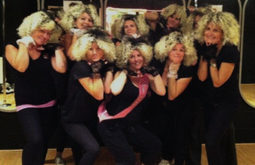 Group of women at a Madonna themed hen party dance class wearing curly wigs and black outfits, posing together in a fun studio setting.