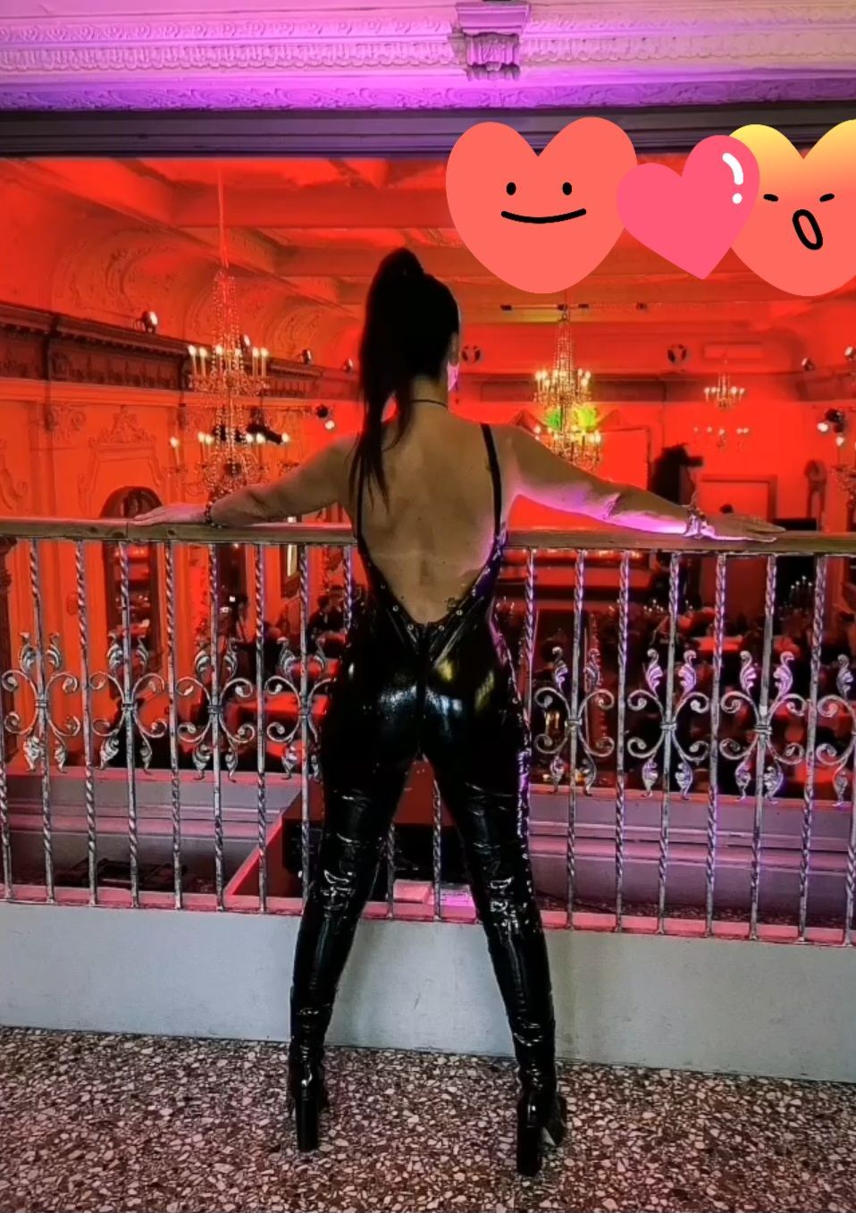Mistress CiCi standing confidently in a black glossy latex catsuit and high heels overlooking a luxury dungeon-style venue with red ambient lighting. Professional dominatrix and fetish hostess available in Cardiff, Bristol and surrounding areas with travel available across the UK & Abroad