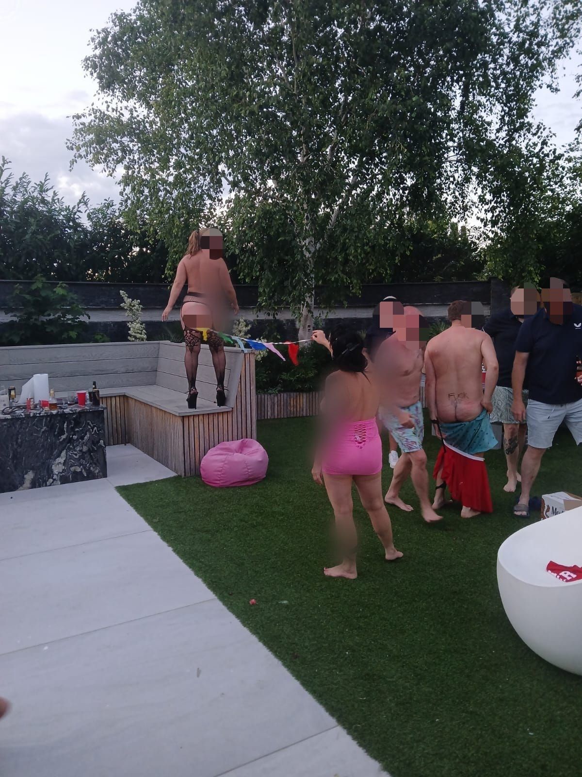 Two female strippers performing an interactive strippergram game at a private party, with one girl pulling colourful bunting from her vagina while the other holds it so guests can play limbo underneath, with Epic Moments UK.