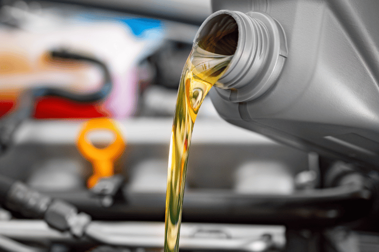 Engine oil pouring from a gray bottle into a car engine compartment, with a blurred orange dipstick in the background