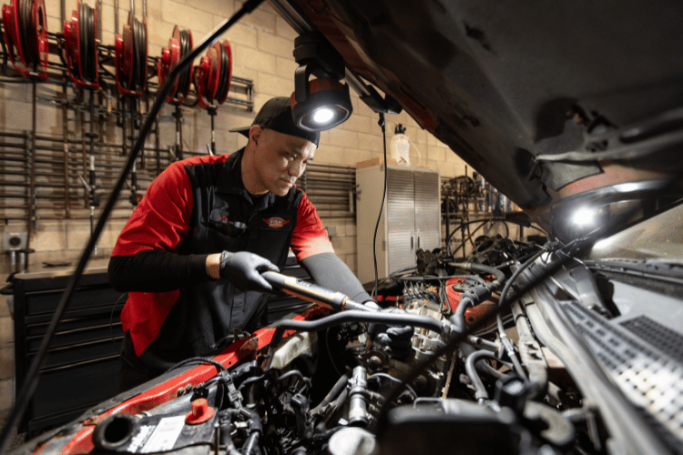 Mechanic in red shirt and black gloves working under an open car hood in a garage