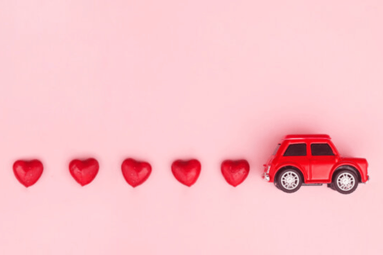 Red toy car driving past a row of red heart-shaped candies on a pink background