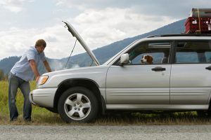 Man leaning over a silver SUV with its hood open on a roadside, mountains in the background