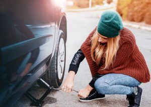 Woman crouching by a car, using a jack to change a tire on a roadside.