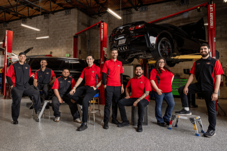 Team of auto mechanics in red shirts posing in a garage beside a lifted black truck.