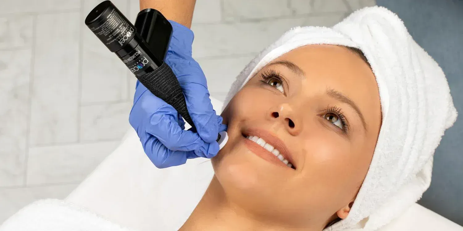 Woman receiving facial treatment, with gloved hand holding a device near her face. She's smiling, lying down.