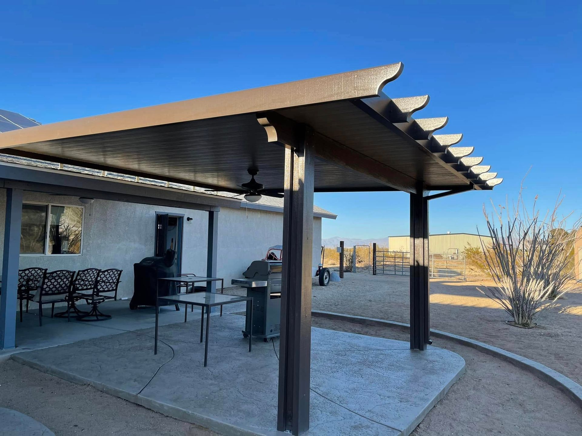 Patio with brown roof and posts, a grill, table, and seating in a desert setting under a blue sky.