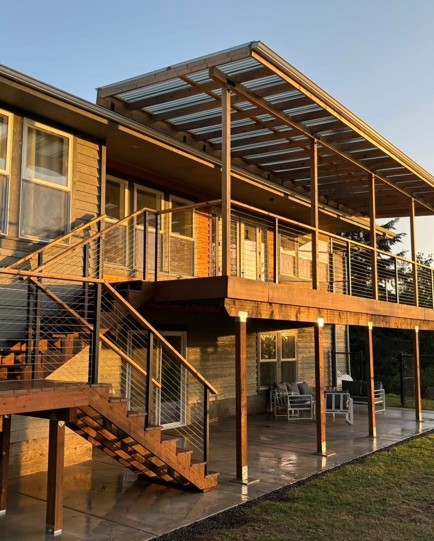 Two-story house with a wooden deck and staircase. Deck has a corrugated roof. Evening sunlight.