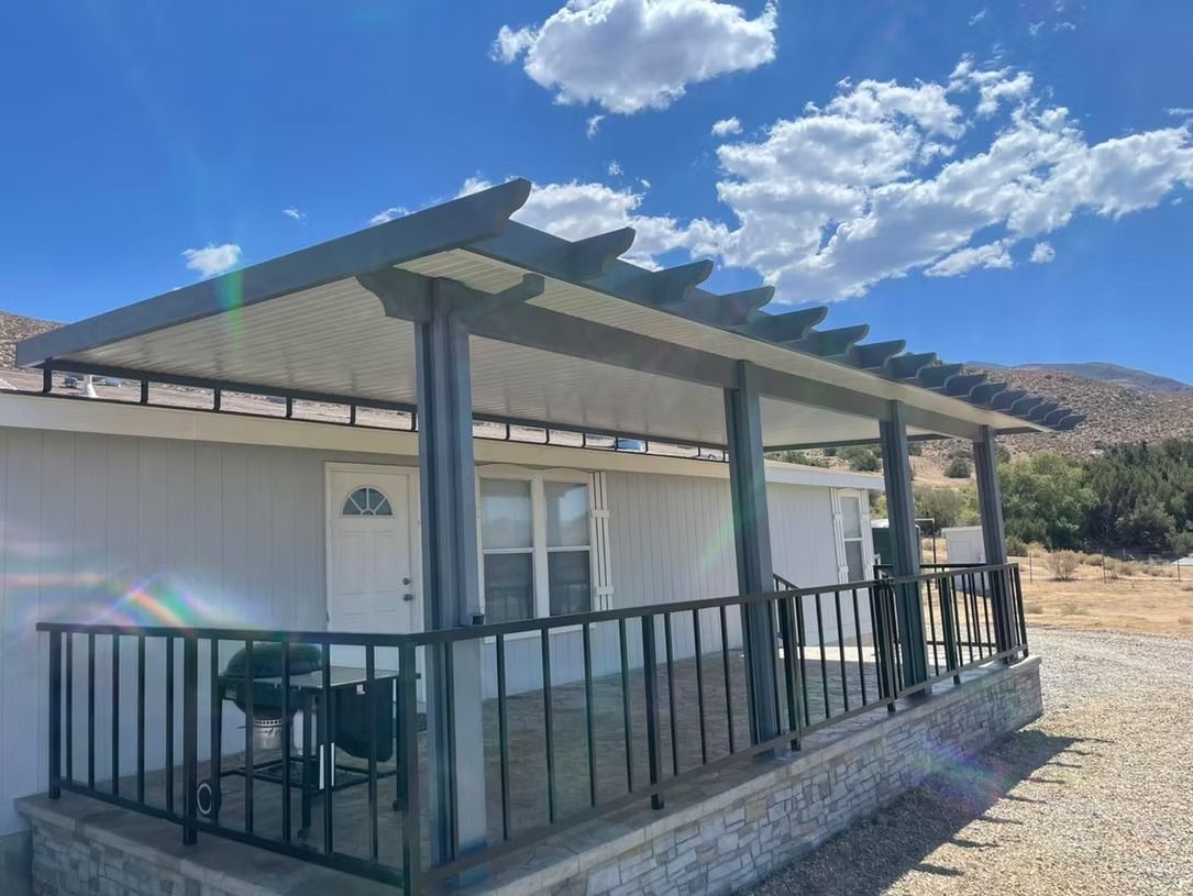 Porch of a white house with a pergola-style roof, black railing, and grill. Blue sky with clouds.