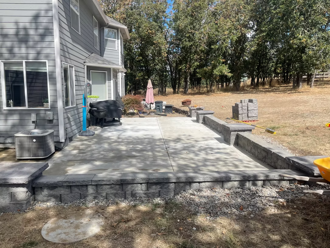 Backyard patio construction with retaining wall and house, with trees and dry grass.