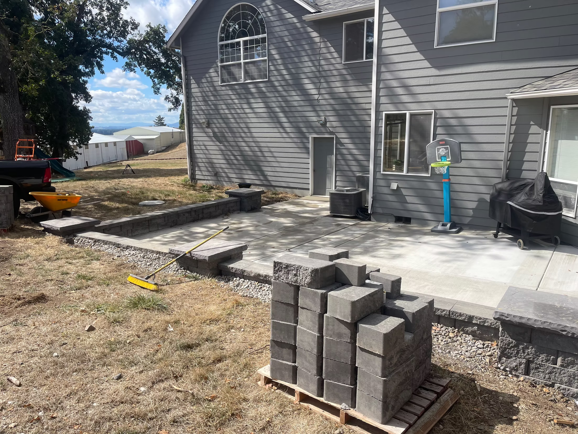 Backyard patio construction in progress; gray pavers, retaining wall, and house.