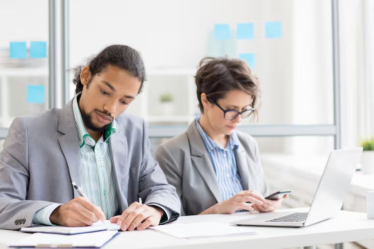 Two people working at a desk: one writing on paper, the other using a phone and laptop in a bright office.