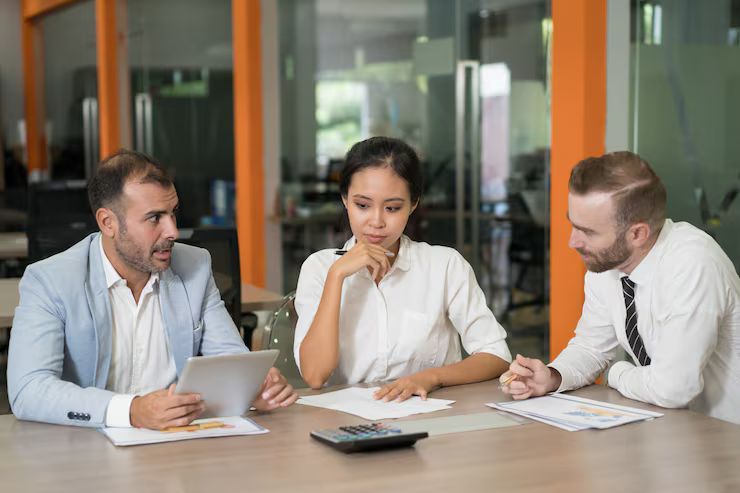 Three people in a meeting, examining papers. One man points at a tablet, others look thoughtful.