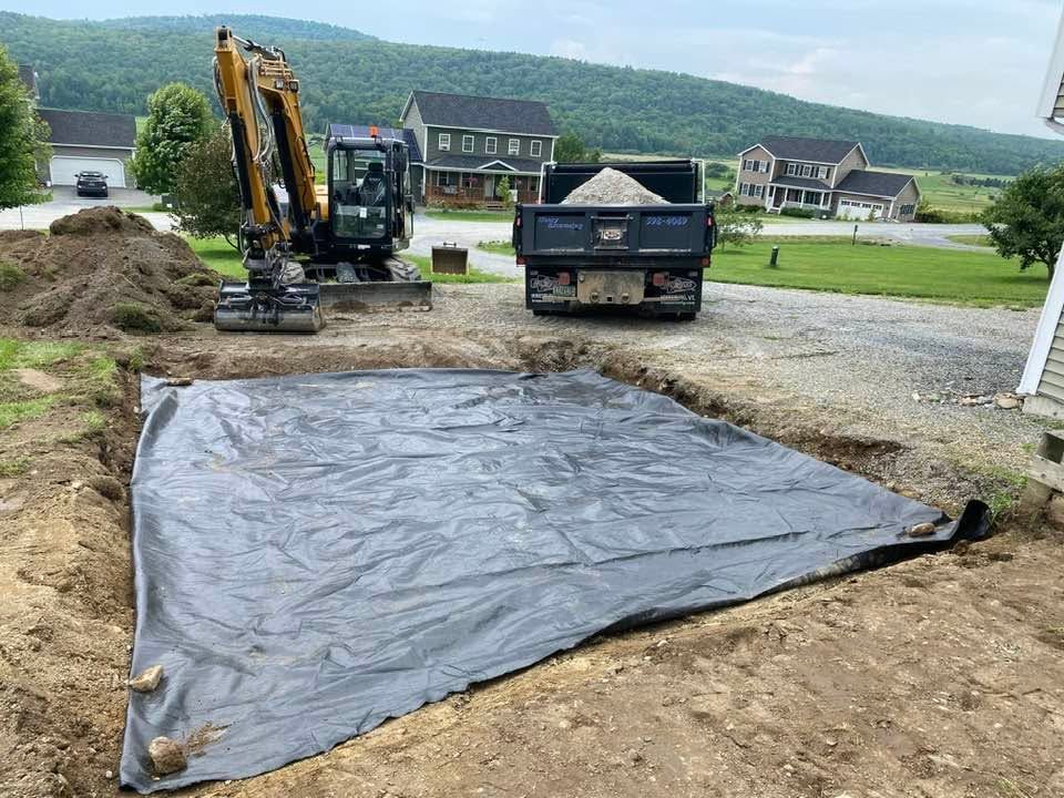 Construction site with excavator and dump truck beside a black tarp-covered pit in a rural area