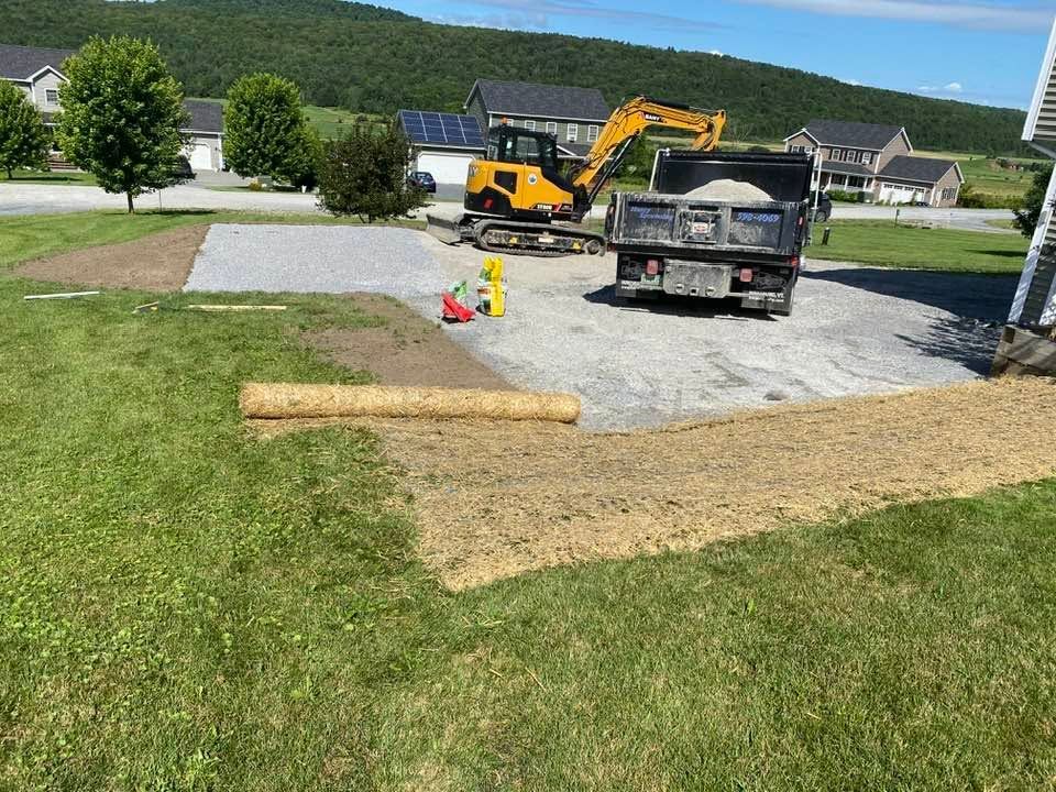 Roadwork with excavator and dump truck on gravel driveway beside a grassy yard and houses