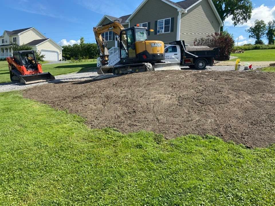 Workers use a mini excavator and truck to spread soil in a suburban front yard.