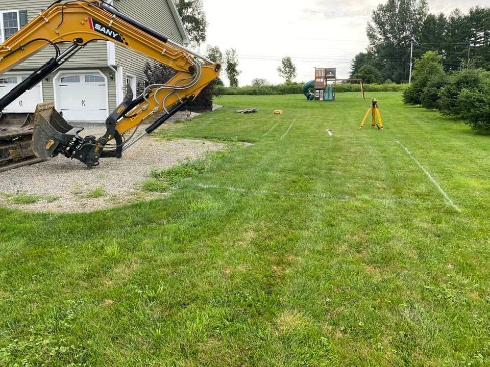 Yellow excavator on a grassy field near a house, with a small orange survey marker in the distance