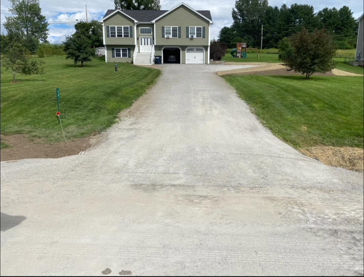 Gravel driveway leading to a gray house with a double garage and front lawn.
