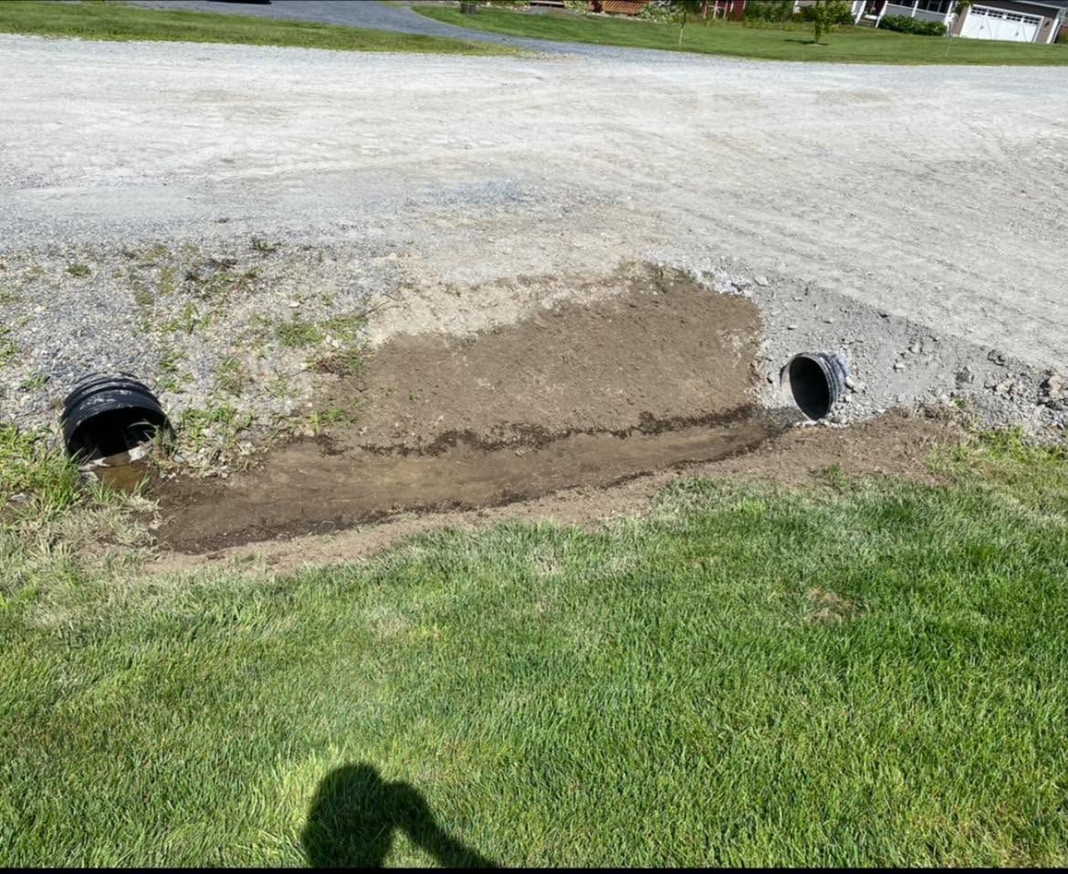 Two culvert pipes under a gravel road beside a grassy ditch, with erosion and muddy soil exposed