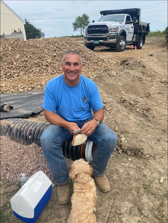 Man sitting on a dirt mound beside a black drain pipe, smiling, with a truck parked in the background.