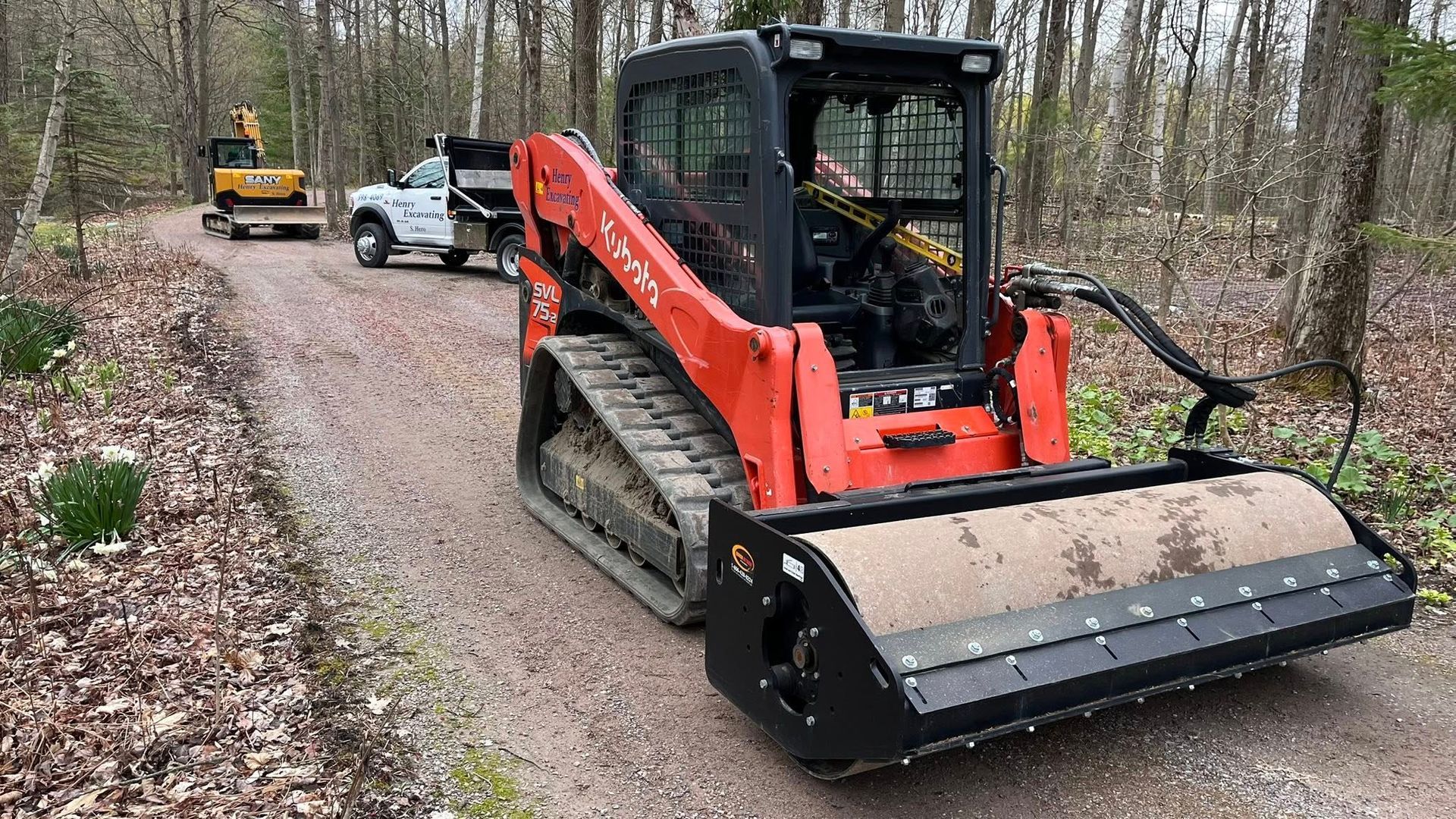 Red skid-steer with brush cutter on a gravel forest road beside a white pickup truck.