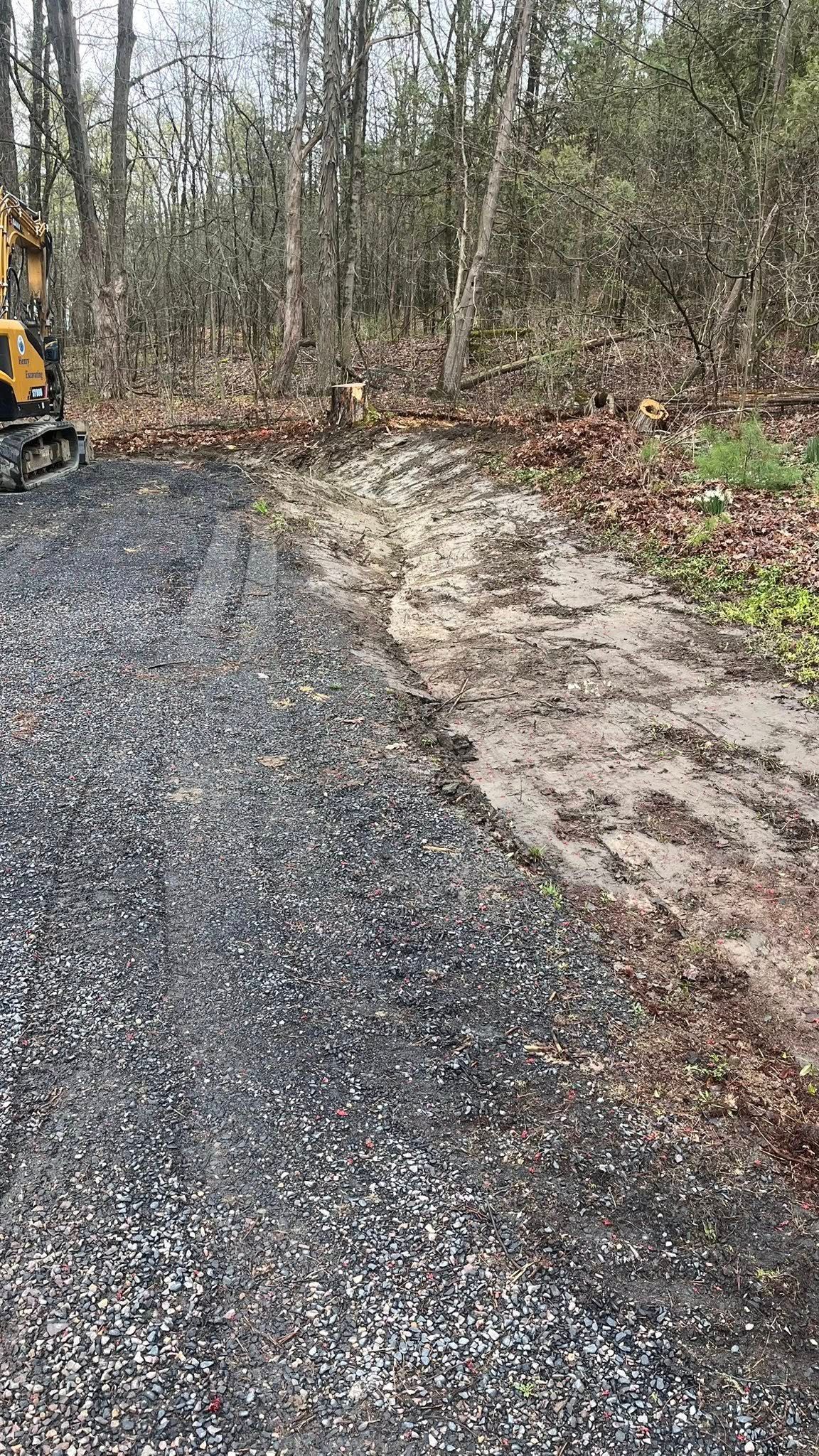 Gravel road beside muddy, leaf-covered edge in a wooded area, with part of a yellow machine at left.
