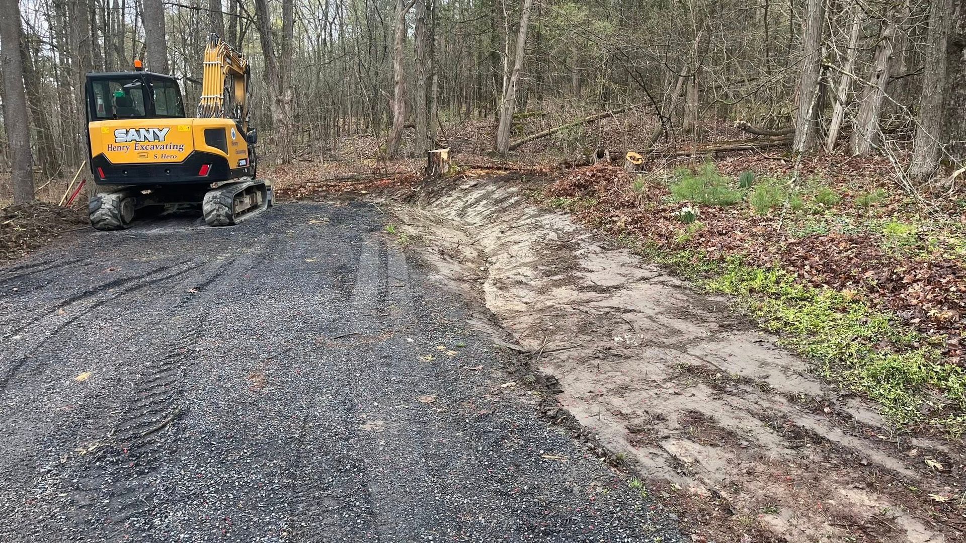 Yellow excavator grading a gravel road beside a muddy wooded slope