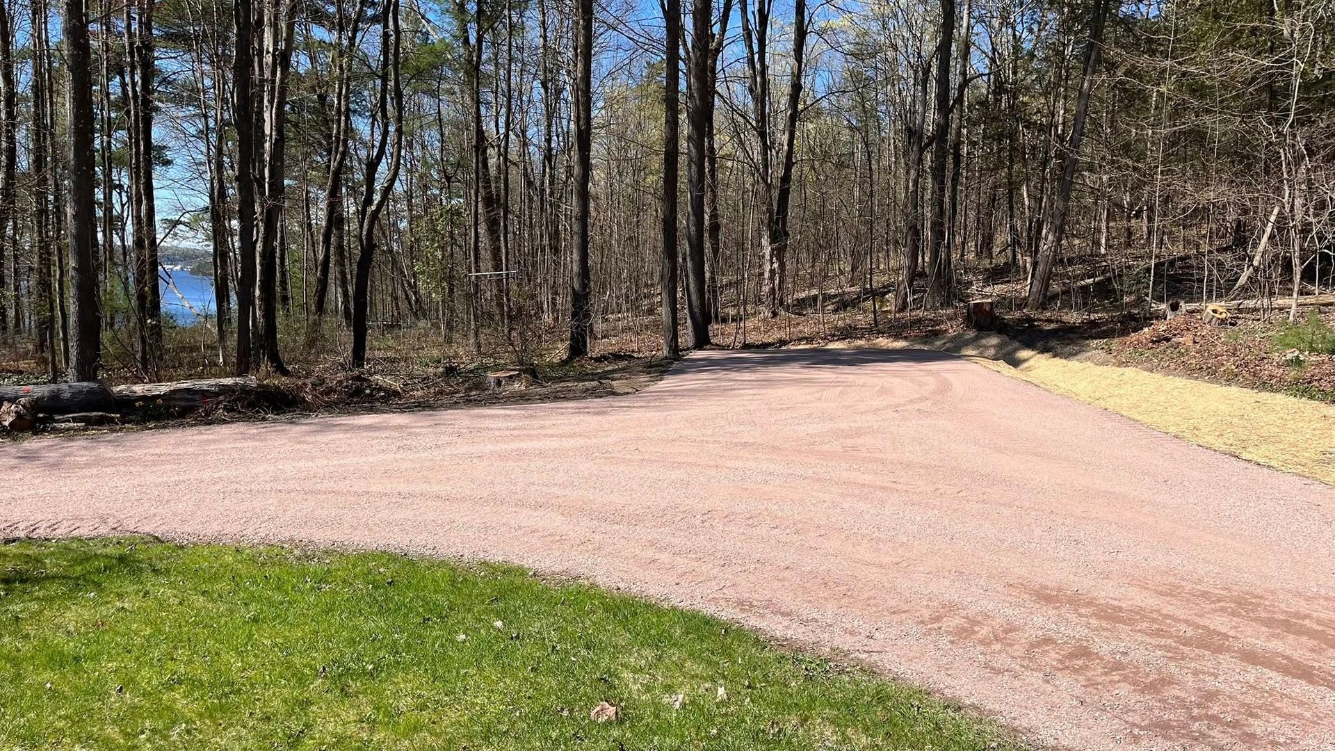Gravel driveway curving through a wooded area with grass in the foreground and water visible through trees