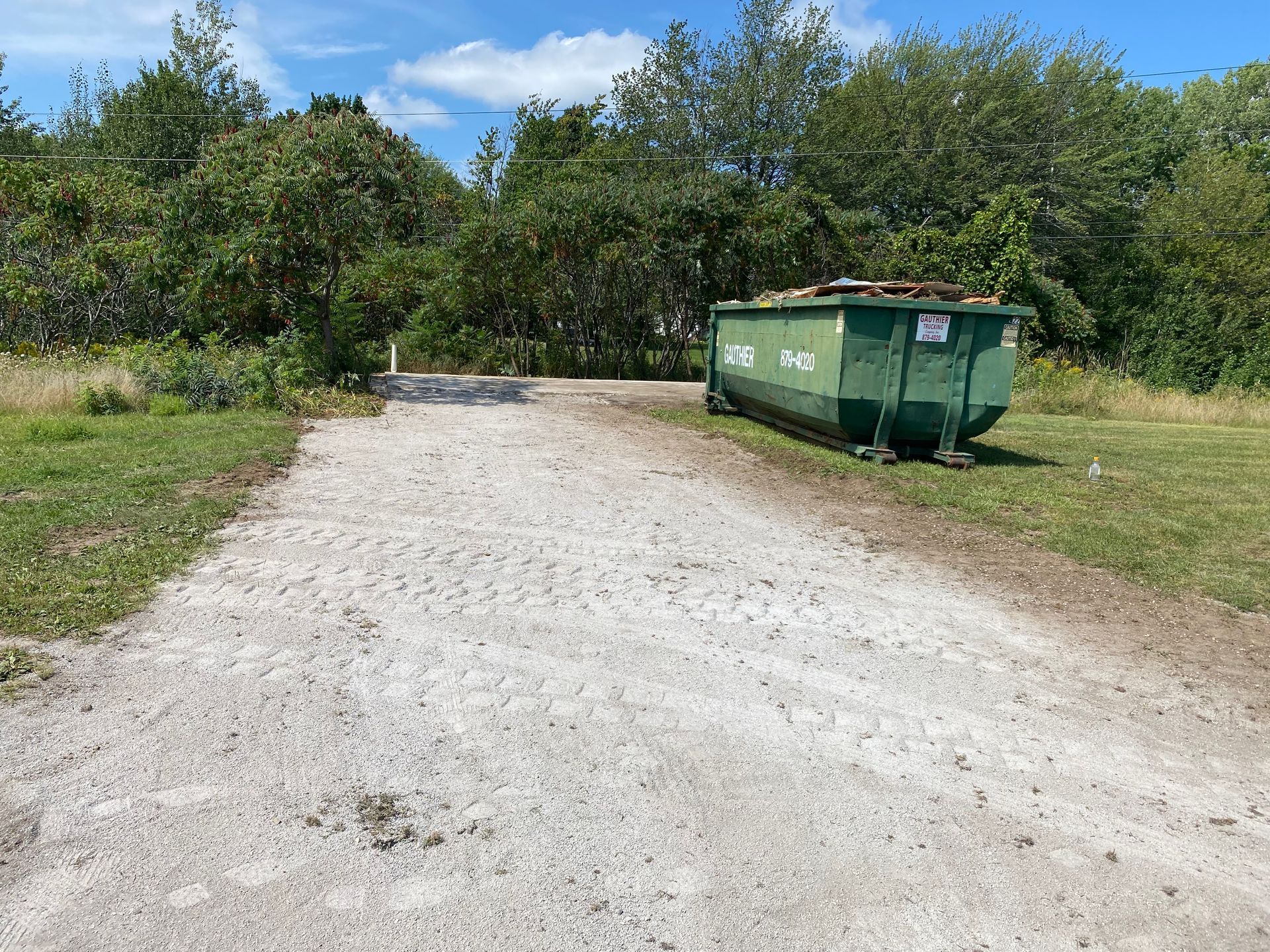 Gravel driveway beside a green dumpster in a sunny, tree-lined area