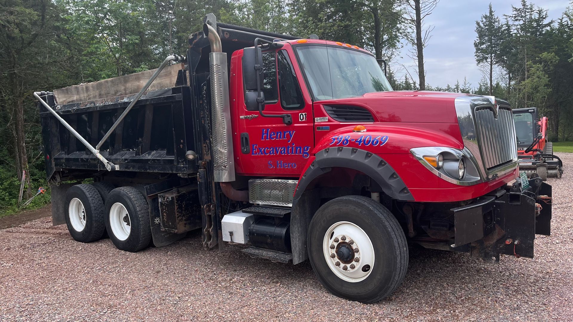 Red dump truck with black bed parked on a gravel lot amid trees.