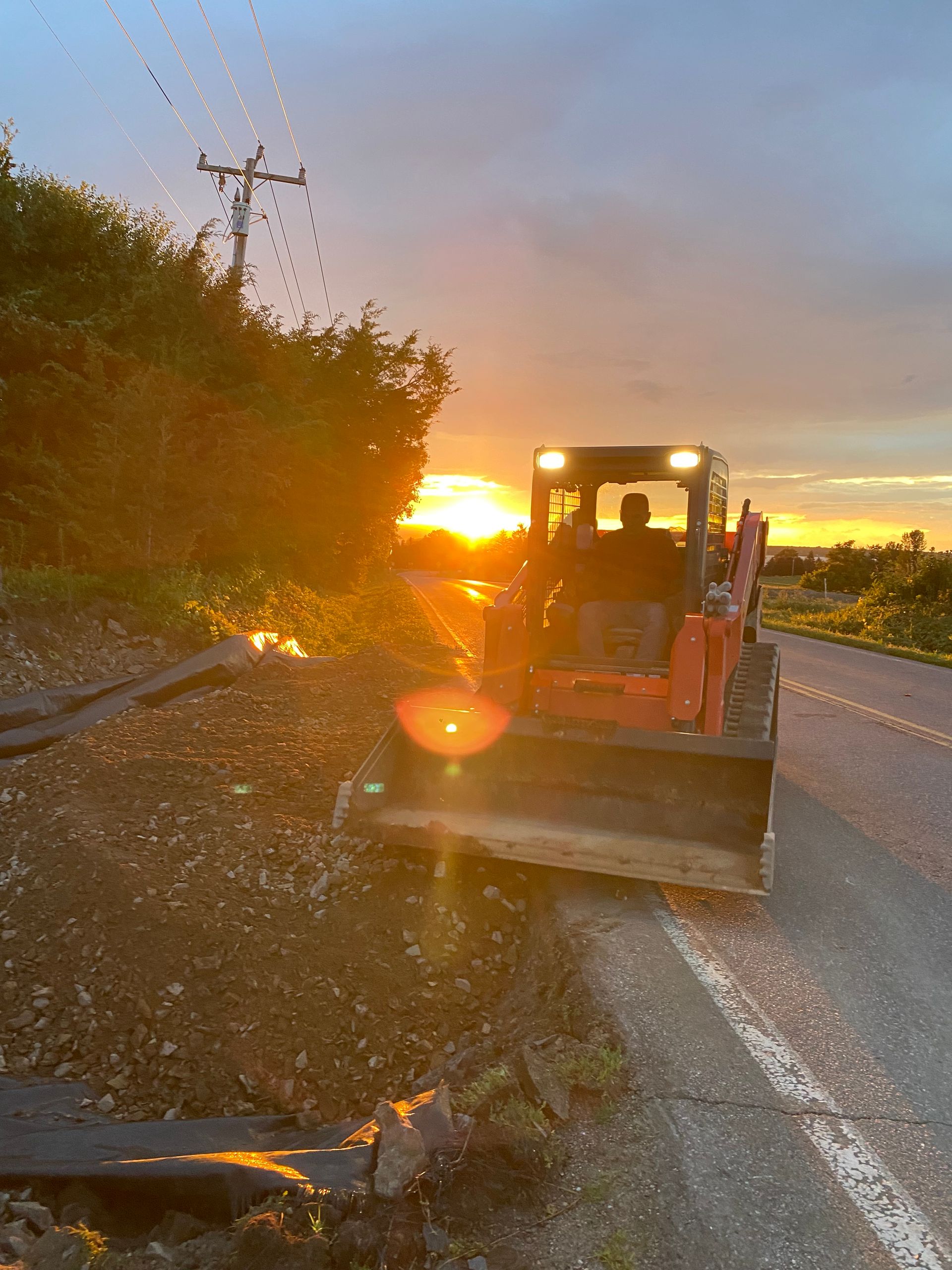 Road grader on a rural road at sunset, with orange light and trees along the roadside.