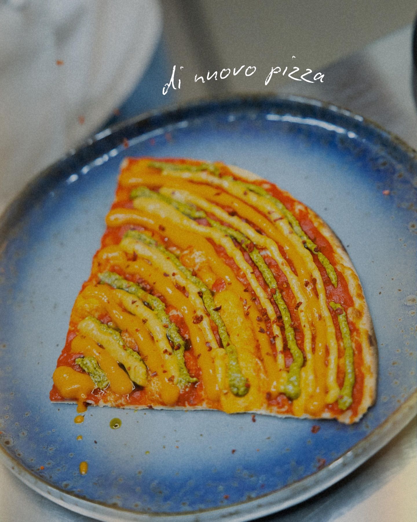 Trancio di pizza su un piatto blu, guarnito con strisce arancioni e verdi. È visibile la scritta