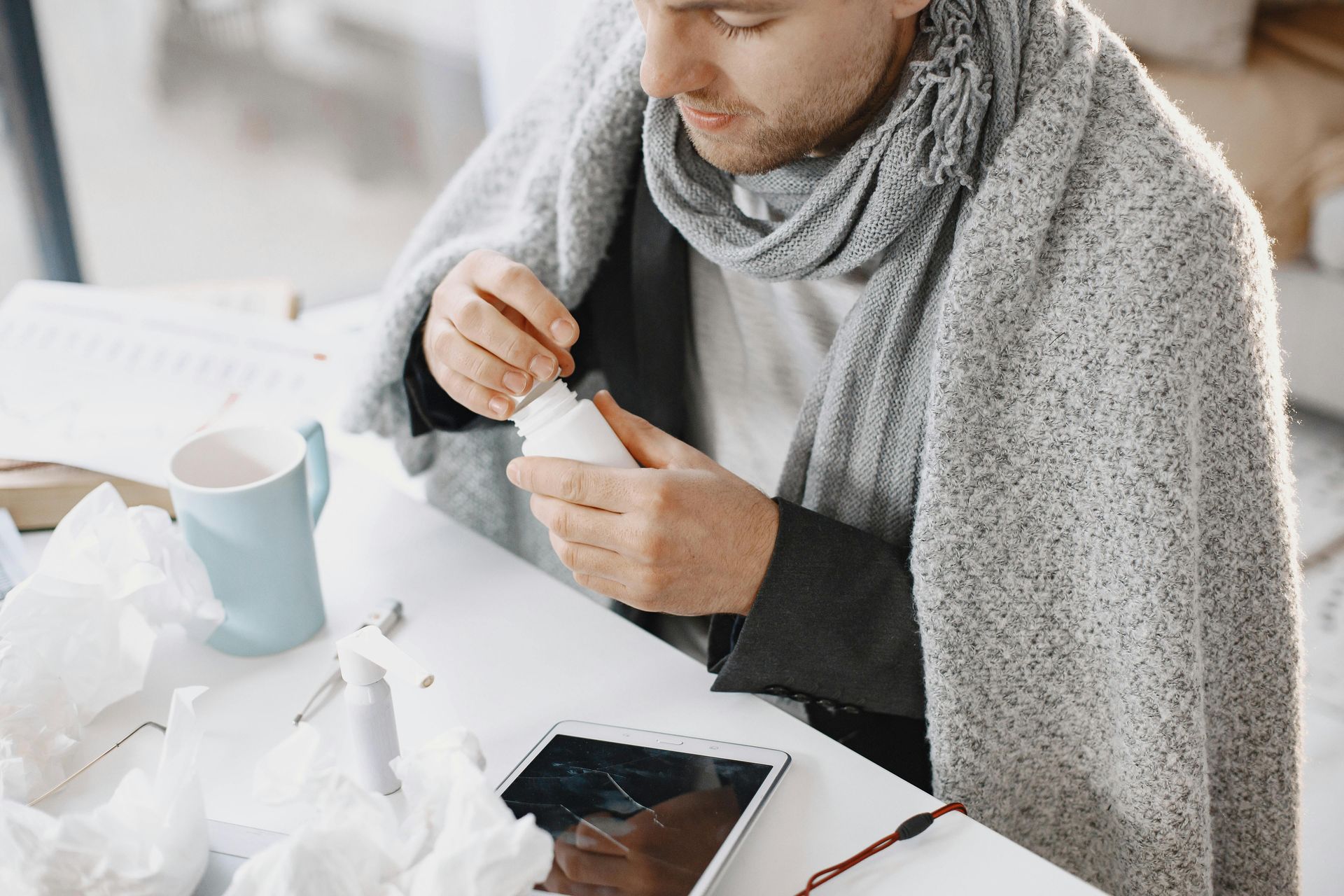 Person in a gray scarf opening a small container at a desk with a mug, tissues, and a tablet