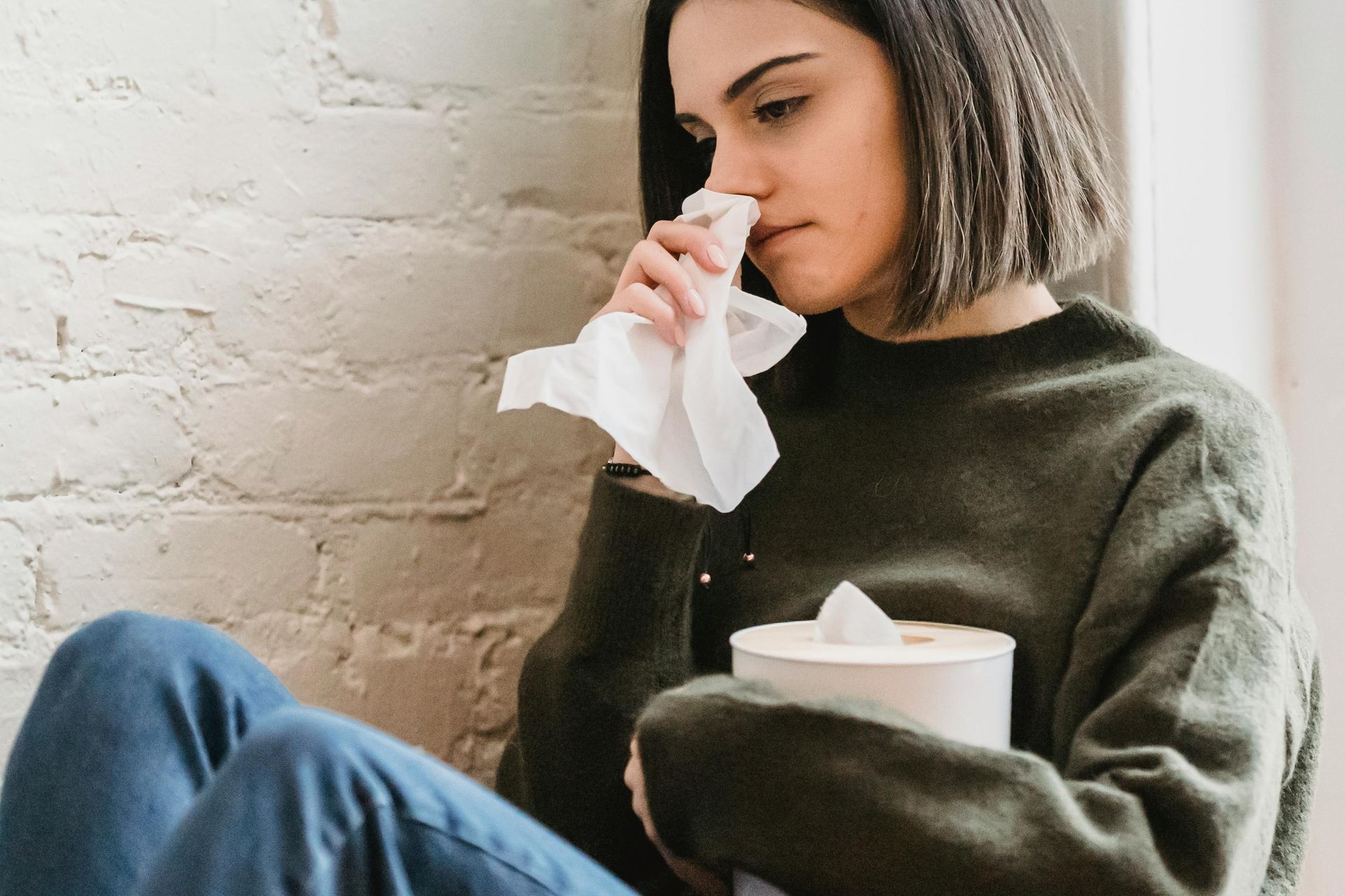 Person sitting on a couch, holding a tissue and tissue box, looking unwell against a white brick wall.