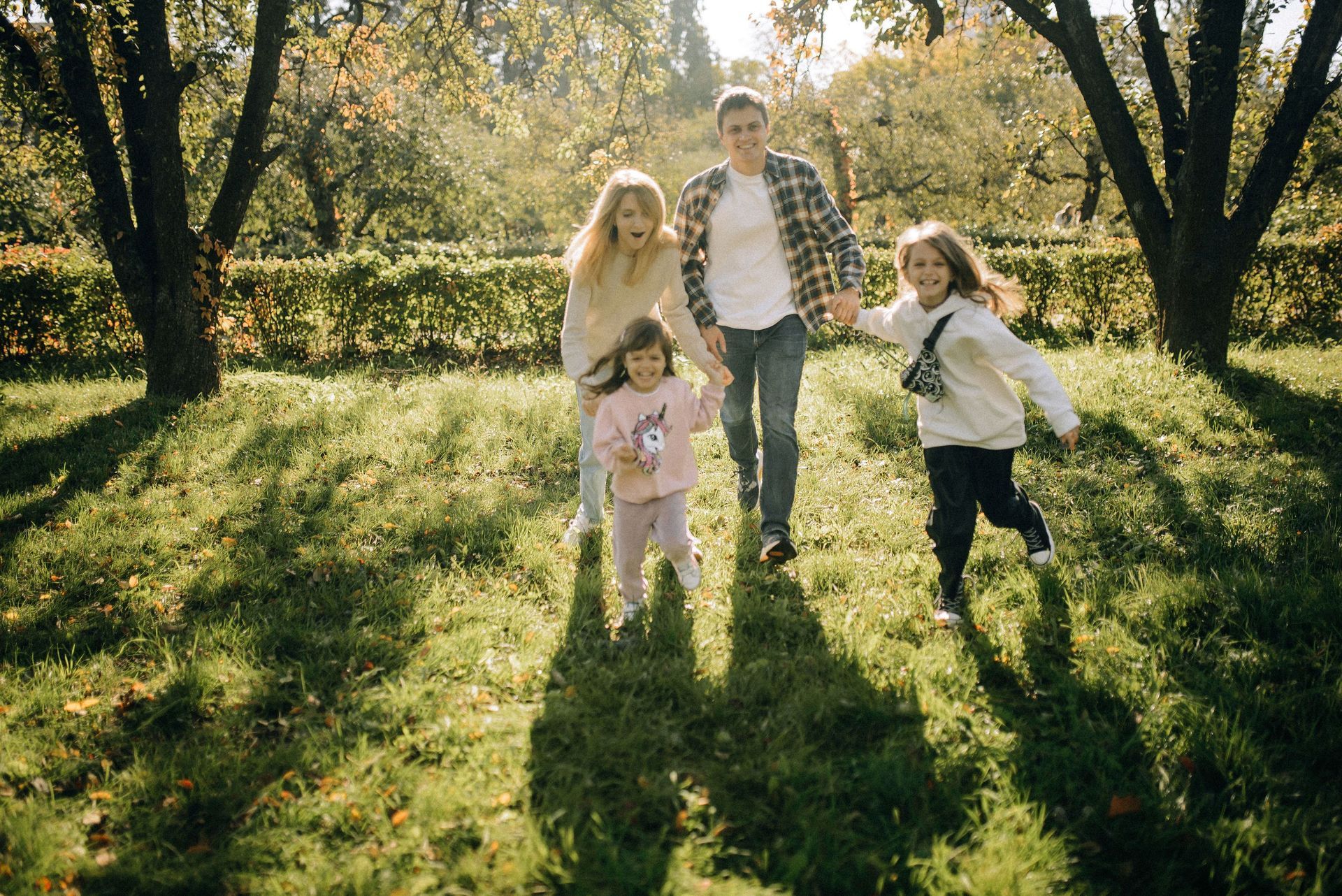 Family of four walking hand in hand across a sunny green lawn framed by tall trees